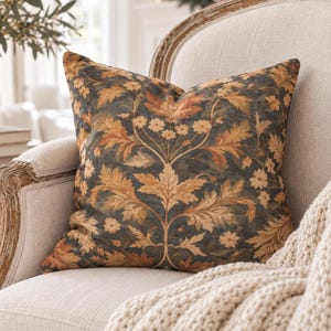 May include: A square throw cushion featuring a dark charcoal background with a symmetrical botanical pattern of golden-brown acanthus leaves and small cream-coloured flowers. The fabric has a textured appearance with intricate floral details.