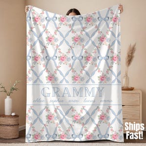May include: White blanket with a floral pattern of pink and blue flowers, light blue bows, and a lattice design. The word "Grammy" is displayed in the center, with names below. A cozy and decorative home textile.
