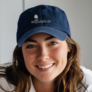 May include: Navy blue baseball cap with the word "an/hophile" embroidered in white, along with a small daisy graphic. The cap is made of a soft material and has a curved brim. A casual accessory for everyday wear.