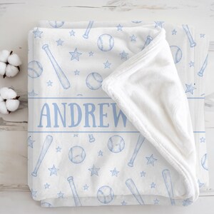 Op de afbeelding: Witte babydeken met een honkbalthema. De deken heeft een patroon van honkballen, knuppels en sterren in lichtblauw. De naam "ANDREW" is in blauw in het midden gedrukt. De deken is opgevouwen en toont een zachte, witte binnenkant.
