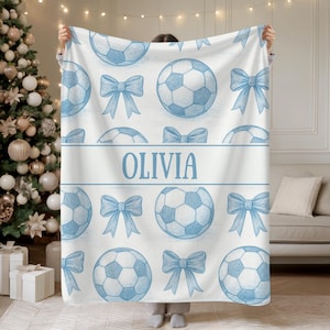 May include: A white blanket featuring a pattern of light blue footballs and bows. The name "OLIVIA" is printed in light blue on a white banner. The blanket is displayed in front of a Christmas tree, with a sofa visible in the background.