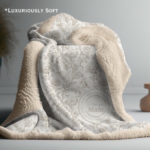 May include: A soft, cream-colored blanket with a floral pattern and a plush, sherpa-like backing. The blanket features a circular design that says "We love you Mom!" and the text "*Luxuriously Soft".