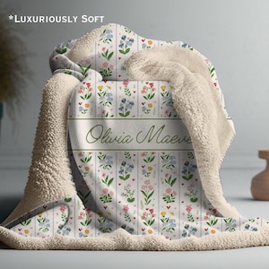 May include: A soft, floral-patterned blanket with a cream-colored sherpa lining. The blanket features a white background with vertical stripes and colorful flowers. The name "Olivia Maeve" is displayed in green script. The text "*Luxuriously Soft" is at the top.