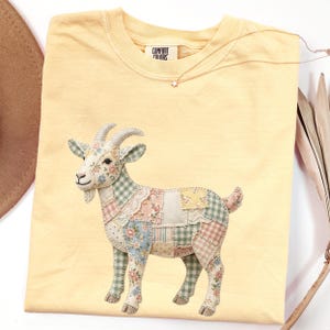 May include: A pale yellow Comfort Colors t-shirt with a patchwork goat design. The goat is decorated with floral and plaid patterns. A star-shaped necklace is visible. The shirt's tag reads "Comfort Colors".