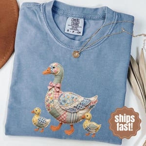 Puede incluir: Camiseta Comfort Colors azul claro con un ganso de patchwork y dos patitos. El ganso tiene un cuerpo con estampado floral, una pajarita rosa y un cuello verde. Los patitos tienen estampados de cuadros amarillos y azules. Un collar de oro está sobre la camisa.