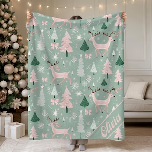 May include: A mint green blanket with a repeating pattern of pink reindeer, Christmas trees, and bows. White snowflakes and trees are also scattered across the blanket. The name "Olivia" is printed in pink on the corner of the blanket.