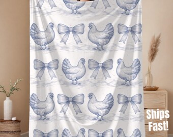 May include: A white blanket featuring a repeating pattern of blue chickens and bows. The chickens are illustrated in a detailed, vintage style, and the bows are tied in a classic shape. The blanket is held up by a person.