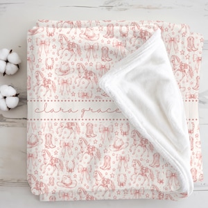 May include: A soft white plush blanket featuring a repeating pattern of red line-art illustrations of horses, cowboy boots, hats, bows, horseshoes, and stars. A horizontal band across the center displays the name Clara Grace in cursive script.