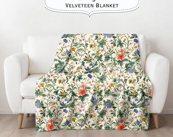 May include: A white velveteen throw blanket with a floral pattern featuring a variety of colorful flowers and leaves. The blanket is draped over a white couch. The text "Chinoiserie Velveteen Blanket" is displayed above the couch.