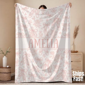 May include: A white blanket with a repeating pattern of pink ballet shoes, bows, and roses. The name "AMELIA" is displayed in pink block letters across the center of the blanket. The blanket is soft and cozy.