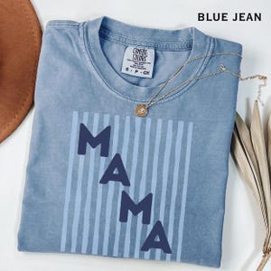Op de afbeelding: Een blauw Comfort Colors T-shirt met het woord "MAMA" in donkerblauwe blokletters. De letters staan tegen een lichtblauwe gestreepte achtergrond. Een gouden ketting met een hanger ligt op het shirt.