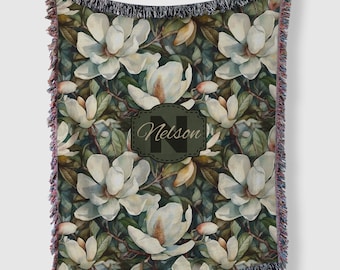 May include: A woven throw blanket with a green and white floral pattern featuring large white magnolia flowers. The blanket has a fringed edge and a personalized label with the name "Nelson" in a black font.