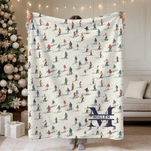 Può includere: Coperta color crema con un motivo ripetuto di sciatori in vari abiti colorati su una pista innevata. La coperta ha un monogramma blu navy con il nome "MILLER" sotto.