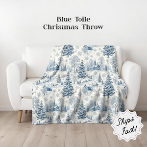 Könnte beinhalten: Eine blau-weiße Weihnachts-Wurfdecke mit Toile-de-Jouy-Design, das Winterszenen mit Häusern, Bäumen und gewundenen Straßen zeigt. Die Decke ist über ein weißes Sofa drapiert.
