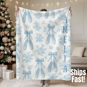 Puede incluir: Una manta blanca con un diseño de zapatillas de ballet azules y flores. El nombre "Amelia" está impreso verticalmente en azul. La manta tiene un aspecto suave y acogedor, ideal para un dormitorio o sala de estar. El texto "Ships Fast!" está en la esquina inferior derecha.
