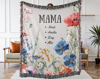 Può includere: Una coperta in tessuto intrecciato con un motivo floreale in tonalità di rosa, blu e rosso. Il testo "MAMA" è stampato in nero nella parte superiore della coperta, seguito da un elenco di nomi: Noah, Amelia, Zoey e Mia.