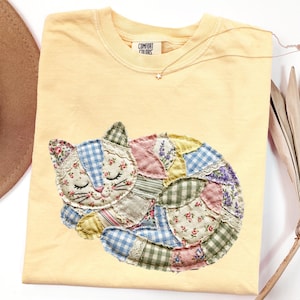 Peut inclure: T-shirt jaune clair Comfort Colors avec un motif de chat en patchwork. Le chat est fait de différents tissus, dont des motifs floraux, vichy et dentelle. Un délicat collier en forme d'étoile est posé sur le t-shirt. Coupe décontractée.