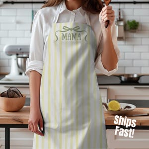 以下が含まれることがあります： 白と黄色のストライプのエプロンで、緑色の文字で「MAMA」と書かれています。エプロンには「MAMA」という言葉の上にリボンのデザインがあります。白いシャツを着た人がエプロンを着用しています。エプロンはキッチンでの使用を目的としています。