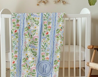 Puede incluir: Una manta floral para bebé con fondo blanco, rayas azules y un círculo monogramado azul con la letra "R". La manta presenta un estampado de flores coloridas y hojas verdes, sobre una cuna blanca.