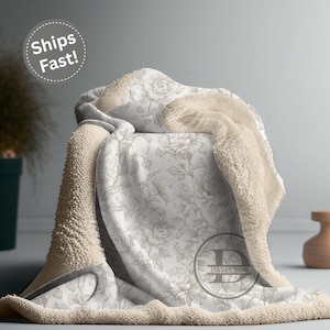 May include: A cozy, floral-patterned blanket with a cream-colored sherpa lining. The blanket features a light beige rose design on a white background. The words "Ships Fast!" are in a white circle in the upper left corner. The blanket is draped over a chair.