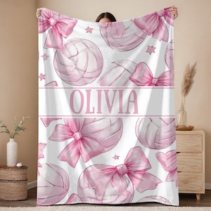 May include: A white blanket featuring a pattern of pink volleyballs, bows, and stars. The name "OLIVIA" is displayed in pink block letters across the center of the blanket. The blanket is soft and cozy.