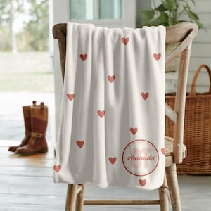 Puede incluir: Una manta color crema con un patrón de corazones rojos está sobre una silla de madera. La manta presenta un diseño circular con el nombre "Amanda" en rojo. La manta está hecha de un material suave.