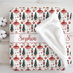 May include: A white plush throw blanket featuring a repeating pattern of red ballerinas, nutcrackers, green Christmas trees, and red ornaments. A horizontal band across the center displays the name Sophia in a red cursive script font.