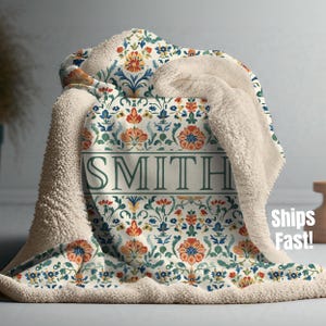 Puede incluir: Una manta de sherpa color crema con un estampado floral en naranja, azul y verde. La manta presenta el nombre "SMITH" en una fuente verde. La manta está doblada, mostrando el suave y esponjoso forro de sherpa.