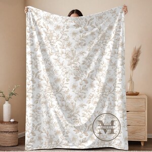 May include: A white blanket with a floral pattern in shades of beige and brown. The blanket features a repeating design of flowers and leaves. A circular emblem with the name "MORRIS" is visible on the lower right corner. The blanket is held up by a person.