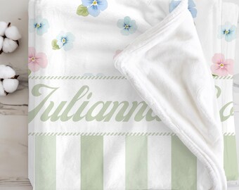 Puede incluir: Una manta de bebé blanca con un estampado floral de flores azules y rosas. La manta tiene un borde a rayas verdes y blancas y el nombre "Julianna" en escritura verde. La manta está doblada, mostrando el interior suave y afelpado.
