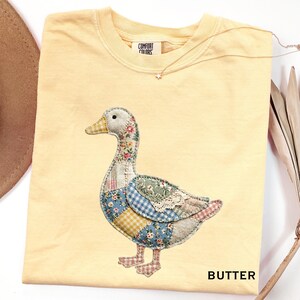 Puede incluir: Una camiseta amarilla clara con un diseño de ganso de patchwork. El ganso presenta patrones florales, de cuadros y de encaje en varios colores. La palabra "BUTTER" está impresa en la parte inferior derecha. También se ve un collar de oro con un colgante de estrella.