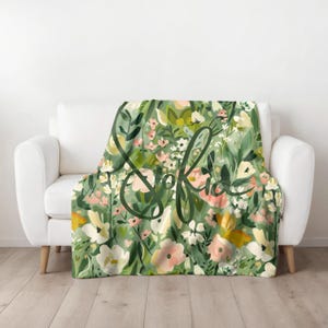 May include: A green floral throw blanket with a white background and the word "Sofia" written in cursive. The blanket features a variety of flowers, including pink, white, and yellow.
