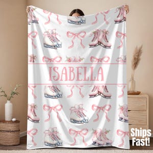 Puede incluir: Manta blanca con un patrón repetido de patines de hielo rosas, lazos y detalles florales. El nombre "ISABELLA" se muestra en rosa en el centro. La manta está sostenida por una persona.