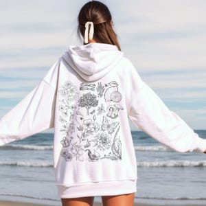Puede incluir: Sudadera con capucha blanca con un estampado botánico en blanco y negro que incluye flores, mariposas, limones y setas.