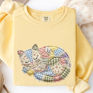 Könnte beinhalten: Hellgelbes Sweatshirt mit einem Patch eines schlafenden Kätzchens. Die Katze besteht aus verschiedenen Stoffmustern, darunter Blumenmuster, Vichy-Karo und Spitze. Das Sweatshirt hat einen Rundhalsausschnitt und lange Ärmel.