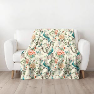 May include: A cream-colored throw blanket with a floral pattern featuring peacocks and pink flowers. The blanket is draped over a white couch.