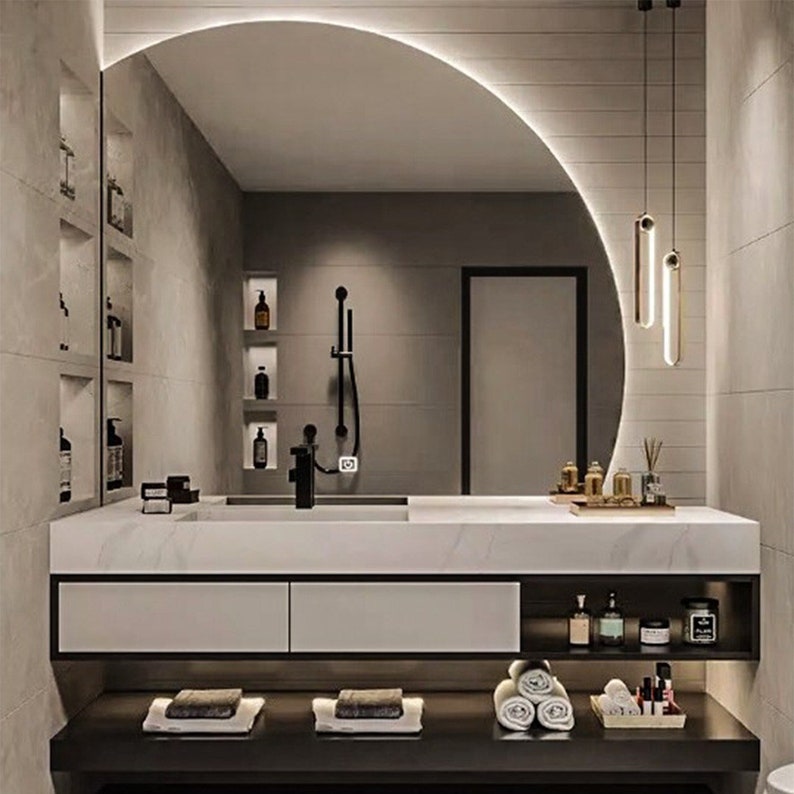 Puede incluir: Un tocador de ba&ntilde;o moderno con una encimera blanca y dos cajones. El tocador tiene un espejo grande y arqueado con una luz incorporada. El ba&ntilde;o tambi&eacute;n cuenta con una ducha con un cabezal de ducha negro y un estante con art&iacute;culos de tocador.