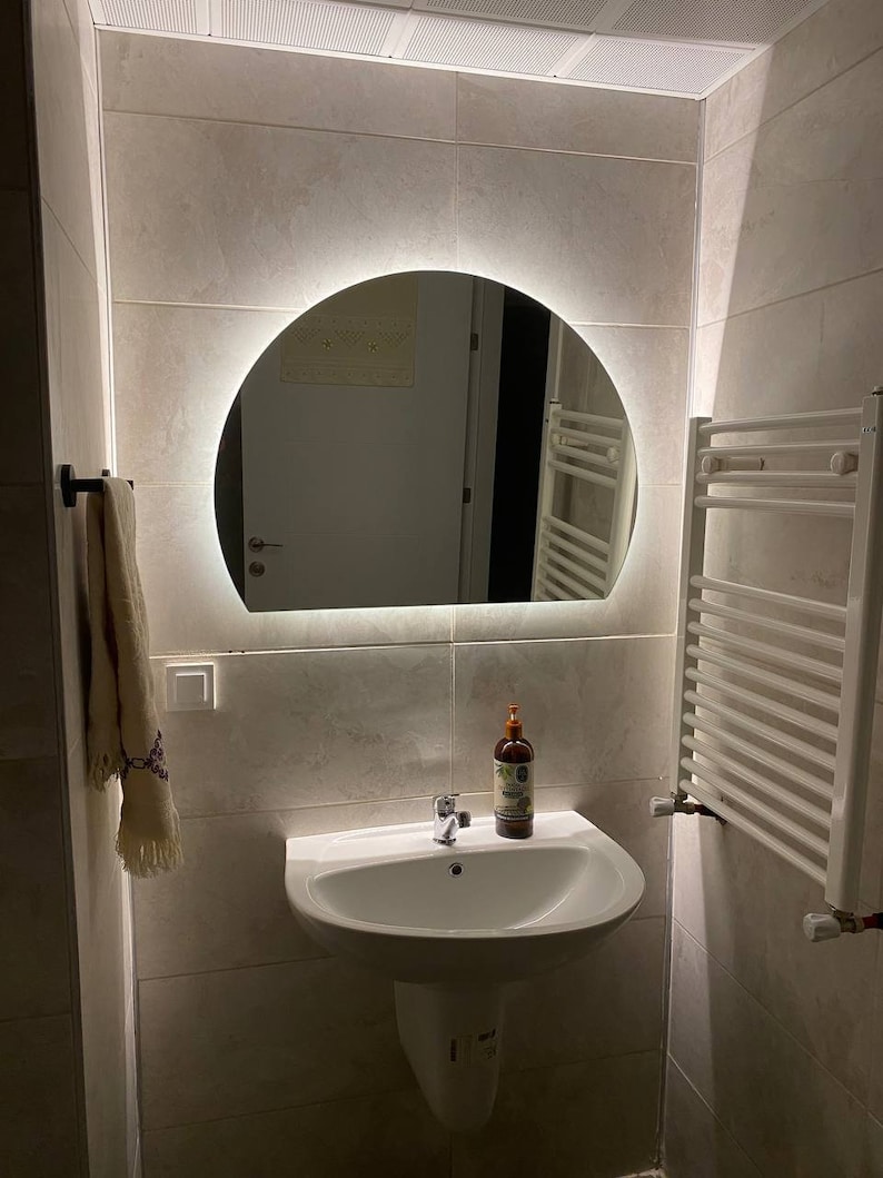 May include: A white bathroom sink with a chrome faucet and a bottle of liquid soap on the counter. The sink is in front of a round mirror with a white light ring around it. A white towel rack is on the right side of the image.