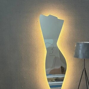 May include: A modern, illuminated mirror with a unique, abstract human figure silhouette. The mirror is lit with warm, white light around the edges, creating a soft glow.