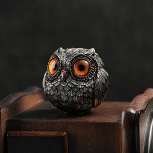 May include: A small, round, silver owl figurine with large, orange eyes. The owl is perched on a wooden surface.