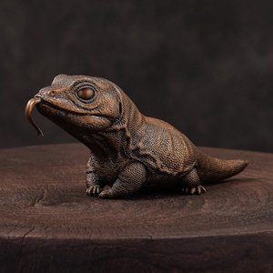 May include: A bronze-colored sculpture of a Komodo dragon with its tongue extended. The reptile is detailed with textured scales and sits on a dark wooden surface. The sculpture is a detailed representation of the animal.