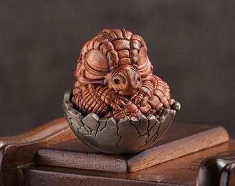 May include: A small, brown, ceramic dragon sculpture curled up inside a cracked, silver egg. The dragon has detailed scales and a closed mouth.