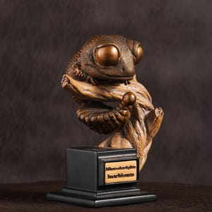 May include: A bronze-colored sculpture of a gecko perched on a wooden branch. The gecko has large, round eyes and textured skin. The sculpture is mounted on a black base with a small plaque that has text on it.