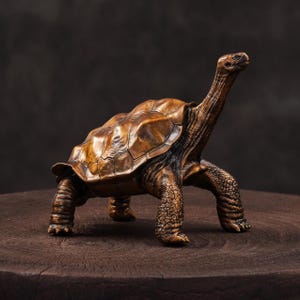 May include: A bronze-colored turtle figurine with a detailed shell and textured skin. The sculpture has a long neck and is posed as if walking. The figurine is displayed on a dark wooden surface.