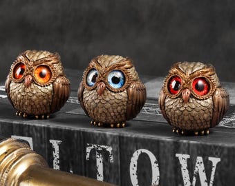 May include: Three decorative owl figurines with detailed feather textures and varying eye colors: orange, blue, and red. The owls are bronze-colored and sit on a book with the word "TOW" visible. A brass telescope is in the foreground.