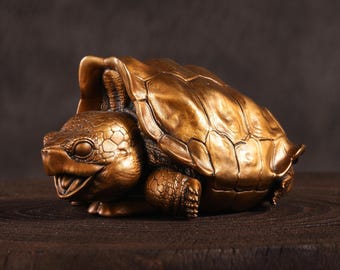 May include: A bronze-colored turtle figurine with a detailed shell and smiling face. The sculpture is resting on a dark wooden surface, showcasing the intricate texture and design of the turtle.