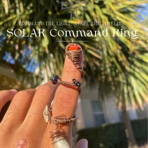 May include: A hand wearing several copper wire-wrapped rings with gemstones. The rings feature a red stone, a blue stone, and a teardrop-shaped white stone. Text on the image reads "COMMAND THE LIGHT. SHAPE THE TIMELINE. SOLAR Command Ring."