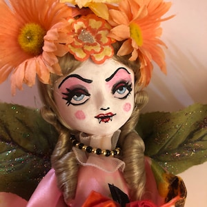 May include: A porcelain doll with blonde hair, wearing a pink dress with orange flowers and a black beaded necklace. The doll has a painted face with blue eyes, pink cheeks, and red lips. The doll is wearing a crown of orange flowers.