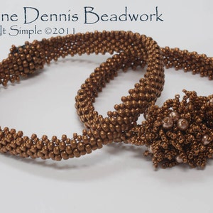 May include: Two brown beaded bracelets with a brown beaded flower charm. The bracelets are made with small, round beads and have a simple, elegant design. The flower charm is made with the same beads and has a delicate, intricate design.