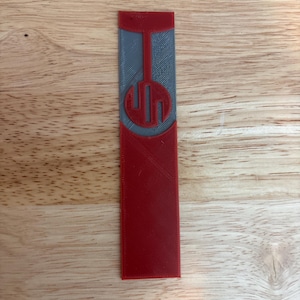 May include: A red and gray 3D-printed bookmark with a geometric design. The bookmark has a rectangular shape with a rounded top and a circular emblem in the center. The emblem features a stylized design within a circle.